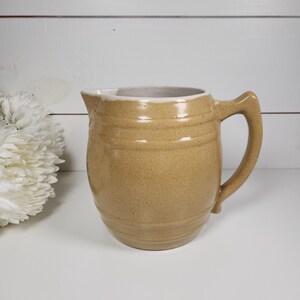 May include: A brown ceramic pitcher with a white rim and a handle. The pitcher has a textured surface and is decorated with horizontal lines.