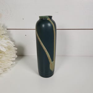 May include: A tall, cylindrical ceramic vase with a matte dark teal finish. The vase features a prominent, light tan, V-shaped decorative glaze accent that runs from the narrow rim down the side, creating a distinct contrast against the dark body.
