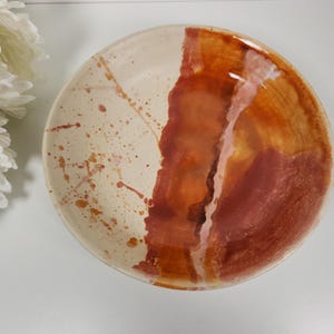 May include: A handmade ceramic plate with a unique design. The plate features a cream-colored base with reddish-brown paint splatters and a vertical band of reddish-brown and orange hues. The plate is round and has a glossy finish.