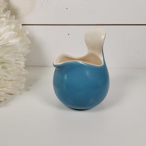 May include: A small, round ceramic creamer with a glossy blue exterior and a smooth cream-colored interior. The vessel features a curved, integrated handle extending upward from the rim and a small pouring spout on the opposite side.