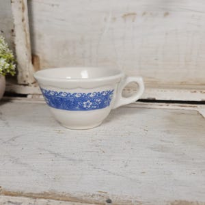 May include: A white ceramic teacup with a blue floral pattern around the rim. The cup has a handle and is sitting on a white wooden surface.