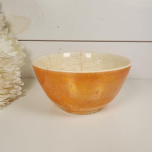 May include: A round ceramic bowl featuring a vibrant orange exterior with a subtle iridescent sheen. The interior of the bowl is a creamy off-white color, showing a network of fine, dark crackle lines across the surface of the glaze.