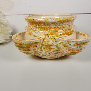 May include: A ceramic serving dish with a speckled pattern in orange, green, and white. The dish has a round top section and three attached, petal-shaped compartments below. The overall design is vintage and decorative.