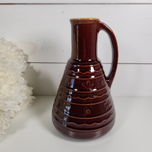 May include: A brown ceramic pitcher with a handle. The pitcher has a wide base that tapers to a narrow neck. It features horizontal bands with floral and circular designs. The rim is a lighter brown color.