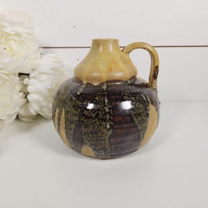 May include: A round ceramic pitcher with a single handle and a narrow neck. The upper portion features a smooth, light tan glaze, while the lower body displays a textured, mottled pattern of deep brown, black, and tan vertical drip-like glaze.