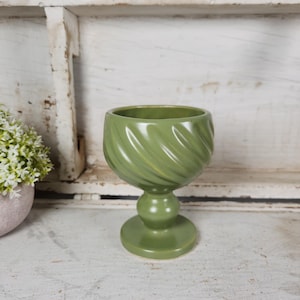 May include: A green ceramic goblet-shaped vase with a pedestal base. The vase has a swirling, textured design on the bowl and a rounded base. The background includes a white shelf and a small pink pot with white flowers.