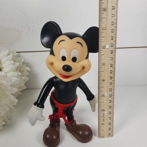 Puede incluir: Una figura vintage de pl&aacute;stico de Mickey Mouse con orejas negras, cuerpo negro, zapatos marrones y una faja roja. La figura mide aproximadamente 18 cm de alto.