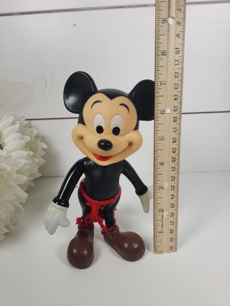 Puede incluir: Una figura vintage de pl&aacute;stico de Mickey Mouse con orejas negras, cuerpo negro, zapatos marrones y una faja roja. La figura mide aproximadamente 18 cm de alto.