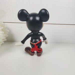 Puede incluir: Una figura de Mickey Mouse de pl&aacute;stico negro con pantalones cortos rojos. La figura est&aacute; de pie sobre una superficie blanca.