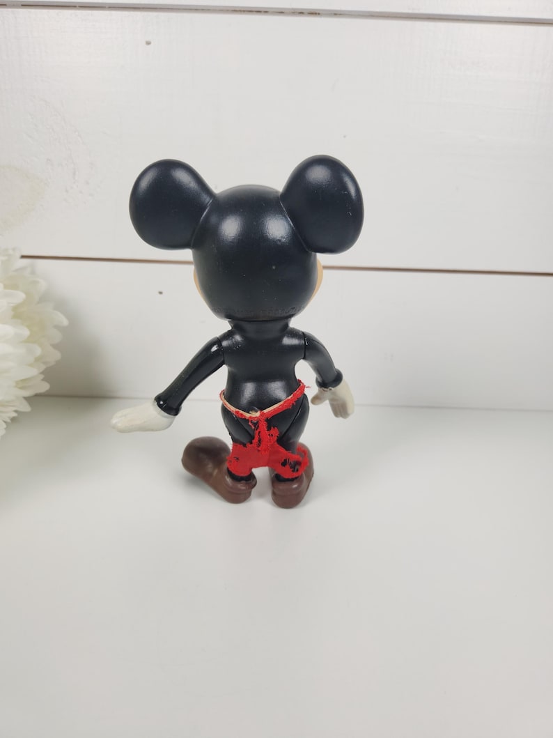 Puede incluir: Una figura de Mickey Mouse de pl&aacute;stico negro con pantalones cortos rojos. La figura est&aacute; de pie sobre una superficie blanca.