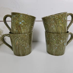 May include: Four olive green ceramic mugs with a speckled pattern. The mugs have a curved handle and a slightly angled rim. The speckled pattern includes shades of blue, brown, and white. The mugs are stacked in pairs.