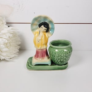 May include: A vintage ceramic figurine featuring a woman holding an umbrella, with a small green pot. The woman wears a yellow robe and a red skirt. The figurine is on a green base.