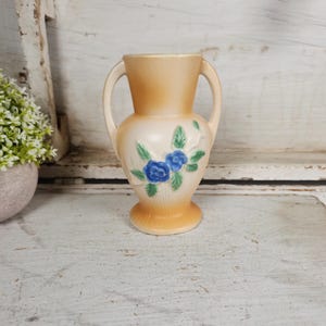 May include: A small, cream-colored ceramic vase with two handles. The vase is decorated with blue and green floral designs.