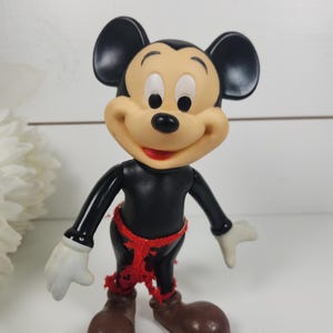 Puede incluir: Una figura vintage de pl&aacute;stico de Mickey Mouse con orejas negras, cuerpo negro, zapatos marrones y un cintur&oacute;n de tela rojo.