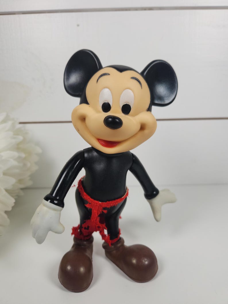 Puede incluir: Una figura vintage de pl&aacute;stico de Mickey Mouse con orejas negras, cuerpo negro, zapatos marrones y un cintur&oacute;n de tela rojo.