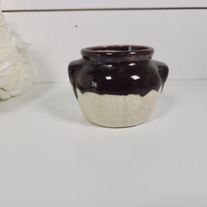 May include: A small, brown and cream-colored ceramic pot with two small handles. The top portion is a glossy, dark brown, while the bottom half is a cracked, cream color. The pot is sitting on a white surface.