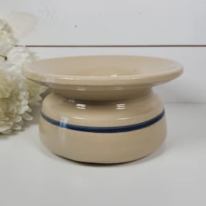 May include: A beige ceramic butter dish with a wide, shallow top and a rounded base. A dark blue stripe encircles the base. The dish has a rustic, vintage aesthetic, suitable for kitchen decor.