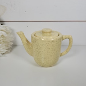 May include: A small, speckled yellow teapot with a matching lid. The teapot has a curved spout and a rounded handle. The ceramic teapot is a vintage style and sits on a white surface.