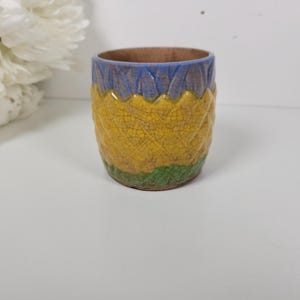 May include: A small ceramic cup featuring a textured, crackled glaze finish. The exterior displays a horizontal color gradient with a blue upper band, a large yellow middle section, and a green base, all with a repeating diamond pattern.