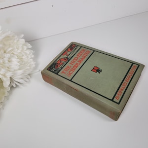 May include: An antique book titled "The Hoosier Schoolmaster" by Edward Eggleston. The book has a green cover with black and red text and illustrations. The book is lying on a white surface.