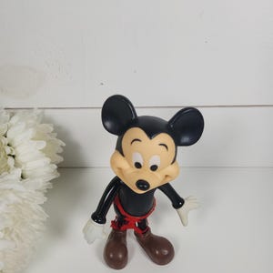 Puede incluir: Una figura de Mickey Mouse de pl&aacute;stico negro y amarillo con pantalones cortos rojos. La figura est&aacute; de pie sobre una superficie blanca.