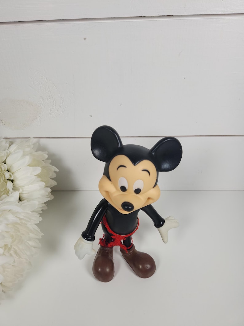 Puede incluir: Una figura de Mickey Mouse de pl&aacute;stico negro y amarillo con pantalones cortos rojos. La figura est&aacute; de pie sobre una superficie blanca.