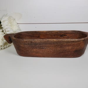 May include: A rectangular, wooden bowl with a dark brown finish. The bowl has two handles on either side. The bowl is sitting on a white surface, with white flowers in the background. This decorative bowl is perfect for home decor.