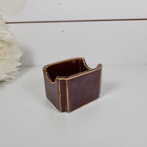 May include: A small, rectangular, brown ceramic container with a curved top and gold trim. The container is sitting on a white surface. The background includes a white flower and a white wooden wall.