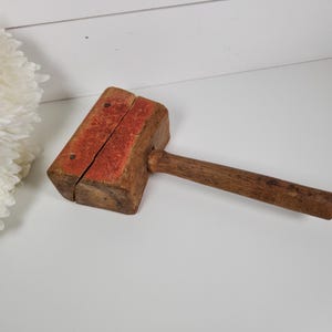 May include: A vintage wooden mallet with a rectangular head and a long, cylindrical handle. The head is painted red on the top and is split down the middle. The mallet is made of a dark brown wood.