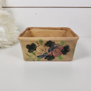 May include: A small, rectangular, beige ceramic container with a floral design. The flowers are painted in shades of pink, blue, and yellow, with green leaves. The container is likely used for holding small items or as a decorative piece.