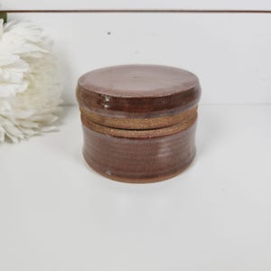 May include: A handcrafted, round ceramic jar with a lid. The jar is a rich, earthy brown color with a textured surface. The lid fits snugly on top, creating a cohesive and functional piece. The jar is suitable for storing small items.