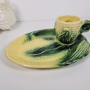 May include: A ceramic corn-themed teacup and saucer set. The cup and saucer are shaped and painted to resemble corn on the cob and leaves. The cup is light yellow with a green handle, and the saucer is yellow and green.