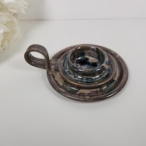 May include: A handmade ceramic candle holder with a handle. The candle holder is a mix of brown, blue, and white colors. The candle holder is round with a handle on the side.