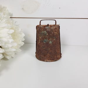 May include: A rustic, weathered metal bell with a handle. The bell has a rectangular shape with a slightly flared base and is covered in rust and verdigris. The bell is likely an antique or vintage item.