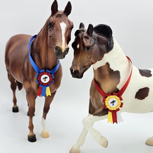 Puede incluir: Dos figuras de caballos, una marrón y otra pía, con cintas de premio. El caballo marrón tiene una cinta azul con detalles rojos, amarillos y azules. El caballo pío tiene una cinta roja con detalles amarillos y blancos.