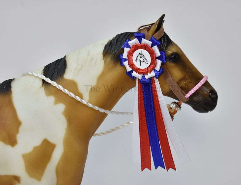以下が含まれることがあります： 茶色と白でペイントされた馬の置物。茶色の革のハルターとピンクのロープのリード、馬の頭が描かれた青、赤、白のリボンを付けている。