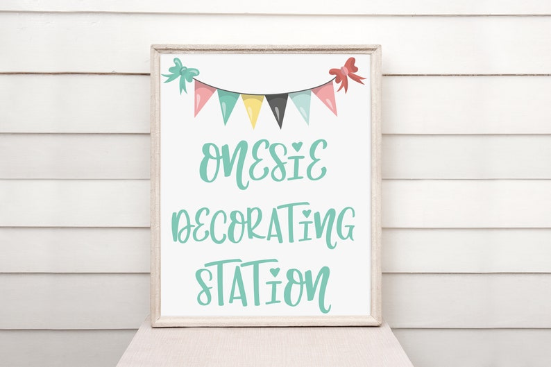 May include: A white sign with a teal frame and a teal banner with a string of colorful flags. The sign says "Onesies Decorating Station" with a heart symbol between each word.