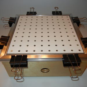 May include: A wooden vacuum forming machine with a white perforated top, secured by black binder clips. The frame is metallic, and the base is light-colored wood. A small hole is visible on the front.