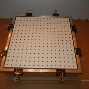 Könnte beinhalten: Eine quadratische, weiße Lochplatte mit gleichmäßig verteilten Löchern, eingerahmt in silberfarbenem Metall. Schwarze Büroklammern befestigen die Platte an einer Holzbasis. Die Lochplatte ist für die Organisation von Werkzeugen oder die Präsentation von Gegenständen konzipiert.