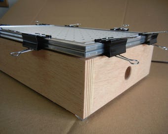 May include: A wooden box with a metal frame and a perforated metal sheet on top. Black binder clips secure the metal sheet to the frame. The box has a circular hole on one side.