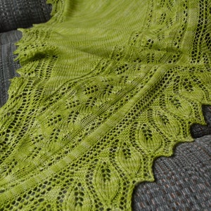 May include: A vibrant green knitted shawl with intricate openwork leaf patterns and a scalloped edge. The shawl is draped over a gray textured surface, showcasing its delicate design and color.