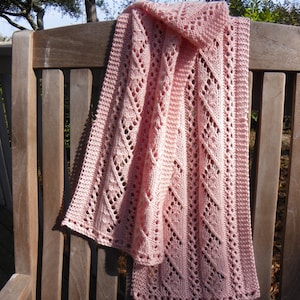 Puede incluir: Una bufanda de punto rosa claro con un diseño de diamantes y flores. La bufanda está sobre un banco de madera. El diseño calado es visible en toda la bufanda, con un borde texturizado. La bufanda está hecha de un material suave y ligero.