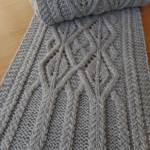 Peut inclure: Une écharpe grise tricotée avec un motif en torsade. L'écharpe est pliée, révélant le motif de tricot détaillé. Fabriquée avec une laine douce et texturée, parfaite pour l'hiver.
