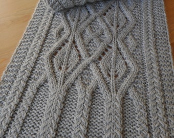 May include: A gray knitted scarf with a cable knit design. The scarf is folded over, showing the intricate pattern of the knit. The scarf is made of a soft, textured yarn, perfect for cold weather.