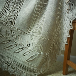 May include: A cream-colored, intricately knitted shawl with a leaf and lace pattern. The shawl is draped over a wooden stool, showcasing the detailed openwork design and scalloped edges. The soft texture and delicate craftsmanship are highlighted.