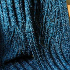 May include: A blue knitted blanket with a cable knit pattern. The blanket is folded and draped over a surface.