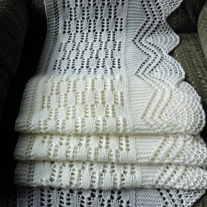May include: A white crocheted shawl with a lacy pattern. The shawl is folded in thirds and is sitting on a sofa.