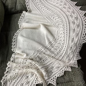 Könnte beinhalten: Ein cremefarbener, gestrickter Schal mit aufwendigen Spitzendetails und einem gewellten Rand. Der Schal ist über einer grauen, gepolsterten Oberfläche drapiert und zeigt sein zartes Lochmuster und seine strukturierte Optik. Das Muster des Schals umfasst Blatt- und wellenartige Motive.