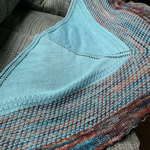 May include: A knitted shawl in shades of teal, blue, and rust. The shawl features a solid teal center with a patterned border. The border has a geometric design with a mix of colors. The shawl is folded on a gray upholstered surface.