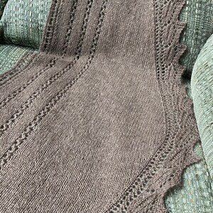 Peut inclure: Un châle en tricot marron avec un délicat motif de dentelle sur les bords. Le châle est drapé sur un canapé vert.